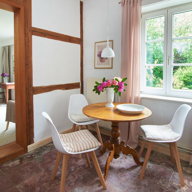 Essbereich in der Künstlerwohnung drei Stühle an einem runden Tisch mit Blumen in einer Vase vor einem Fenster mit rosa Gardinen